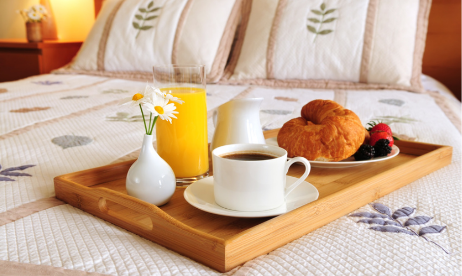 bandeja de café da manhã em cima de uma cama, com jarra de suco, café, xicara, prato com pães, frutas e um vaso com flores.
